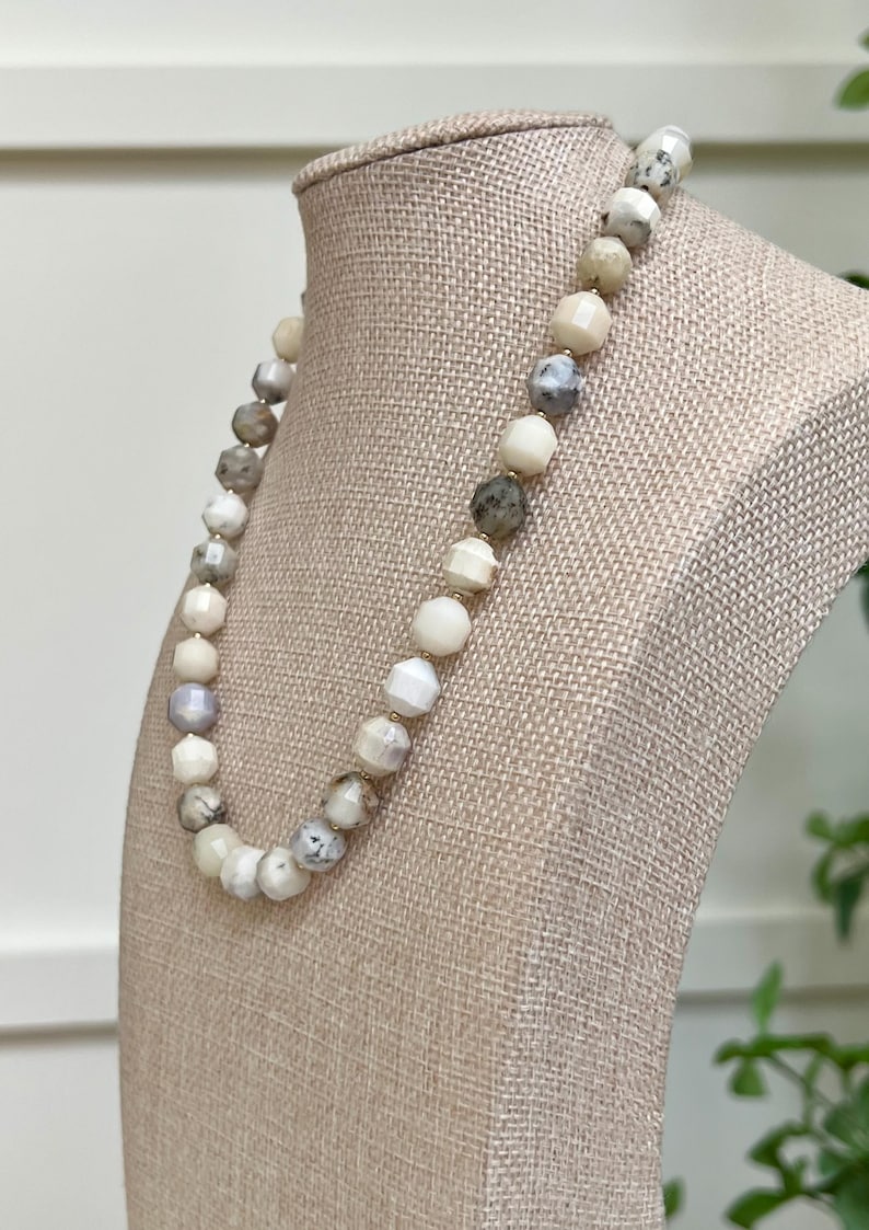 May include: A beaded necklace with a variety of neutral-toned beads, including white, gray, and cream colors. The beads are round and faceted, strung together with gold-colored spacers. The necklace is displayed on a beige mannequin.
