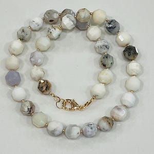 May include: A faceted gemstone necklace featuring a variety of colors including white, gray, and lavender. The necklace has a gold-colored clasp and small gold-colored beads between each gemstone. The gemstones are approximately 0.5 inches in diameter.