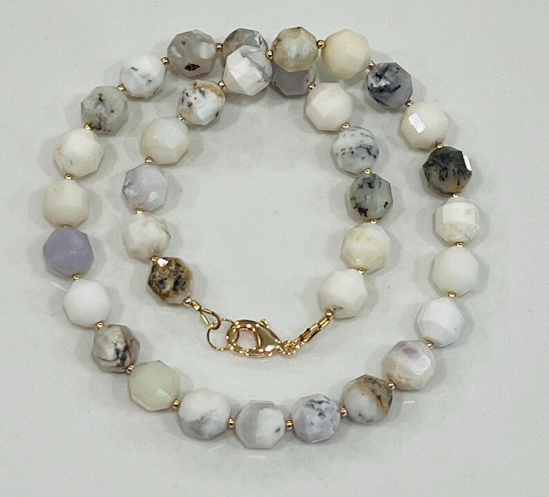May include: A faceted gemstone necklace featuring a variety of colors including white, gray, and lavender. The necklace has a gold-colored clasp and small gold-colored beads between each gemstone. The gemstones are approximately 0.5 inches in diameter.