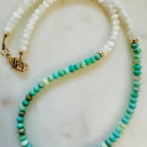 May include: A long necklace with white and turquoise beads. The white beads are on the top half of the necklace and the turquoise beads are on the bottom half. The necklace is made of natural stones and has a gold clasp.