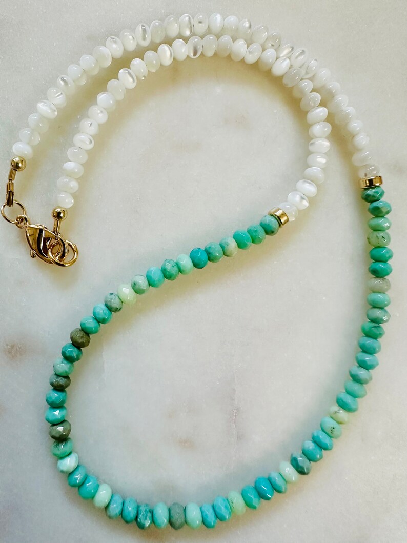 May include: A long necklace with white and turquoise beads. The white beads are on the top half of the necklace and the turquoise beads are on the bottom half. The necklace is made of natural stones and has a gold clasp.