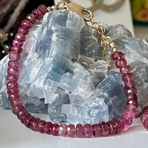 May include: A delicate bracelet featuring faceted, deep pink gemstones strung together with a gold-colored chain and clasp. The bracelet is displayed against a backdrop of a light blue crystal formation, highlighting the jewelry's color and texture.