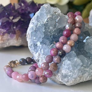 May include: A beaded bracelet with faceted, round gemstones in shades of pink, purple, and brown. The bracelet is draped over a light blue crystal formation. A purple crystal cluster is in the background. The bracelet has a gold clasp.