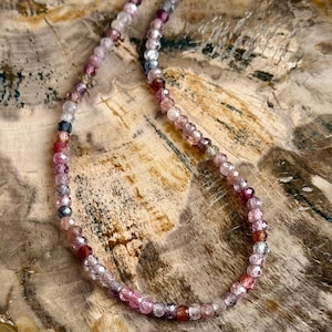 May include: A delicate necklace composed of small, faceted beads in shades of pink, purple, and clear. The beads are strung together to form a necklace. The necklace is displayed on a wooden surface.