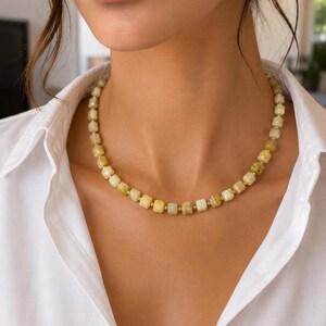 May include: A beaded necklace featuring square-cut, pale yellow gemstones. The necklace is strung with small gold-colored beads between each gemstone. The necklace is worn over a white collared shirt.
