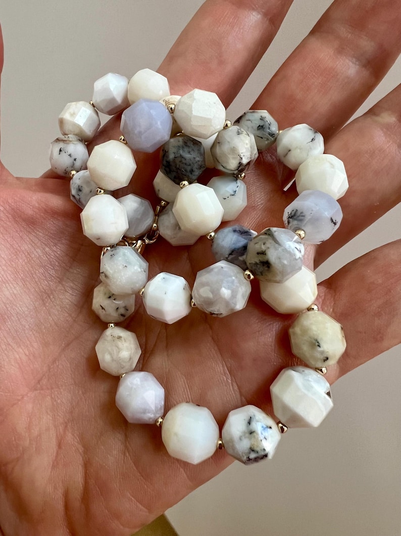 May include: A beaded necklace featuring faceted, irregular-shaped beads in shades of white, gray, and pale blue. The beads are strung together with gold-colored connectors, creating a delicate and elegant piece of jewelry.