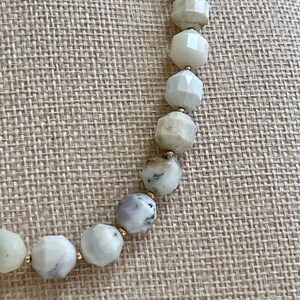 May include: A beaded necklace featuring faceted, light-colored stones with gold-colored spacers. The stones vary in color, including white, gray, and pale green. The necklace is displayed on a neutral-toned fabric.