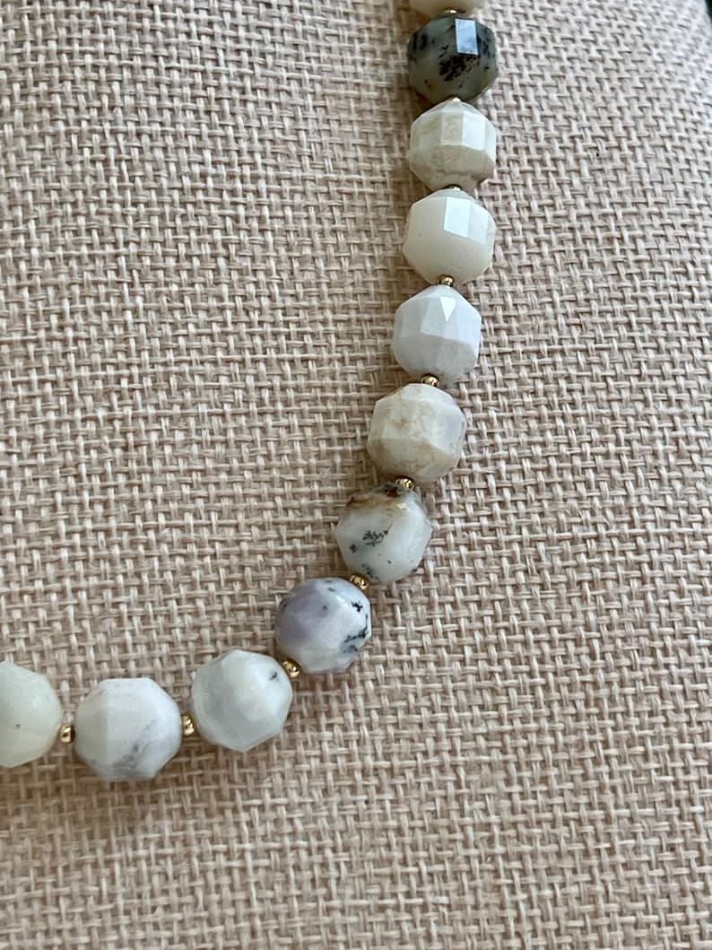 May include: A beaded necklace featuring faceted, light-colored stones with gold-colored spacers. The stones vary in color, including white, gray, and pale green. The necklace is displayed on a neutral-toned fabric.