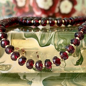 May include: A necklace featuring faceted, deep red garnet beads and gold-coloured spacers. The beads are strung together, forming a circular shape. The necklace is displayed on a reflective surface with a blurred floral background.