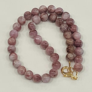 May include: A necklace featuring round, light purple beads with darker inclusions. The beads are strung together, and the necklace is secured with a gold-colored clasp. The jewelry is displayed on a white surface.