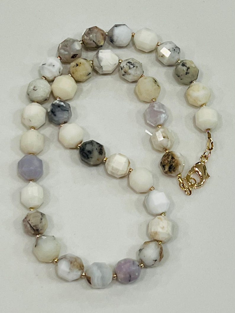 May include: A faceted gemstone necklace featuring a variety of colors including white, gray, and lavender. The necklace has a gold-colored clasp and small gold-colored beads between each gemstone. The gemstones are approximately 0.5 inches in diameter.