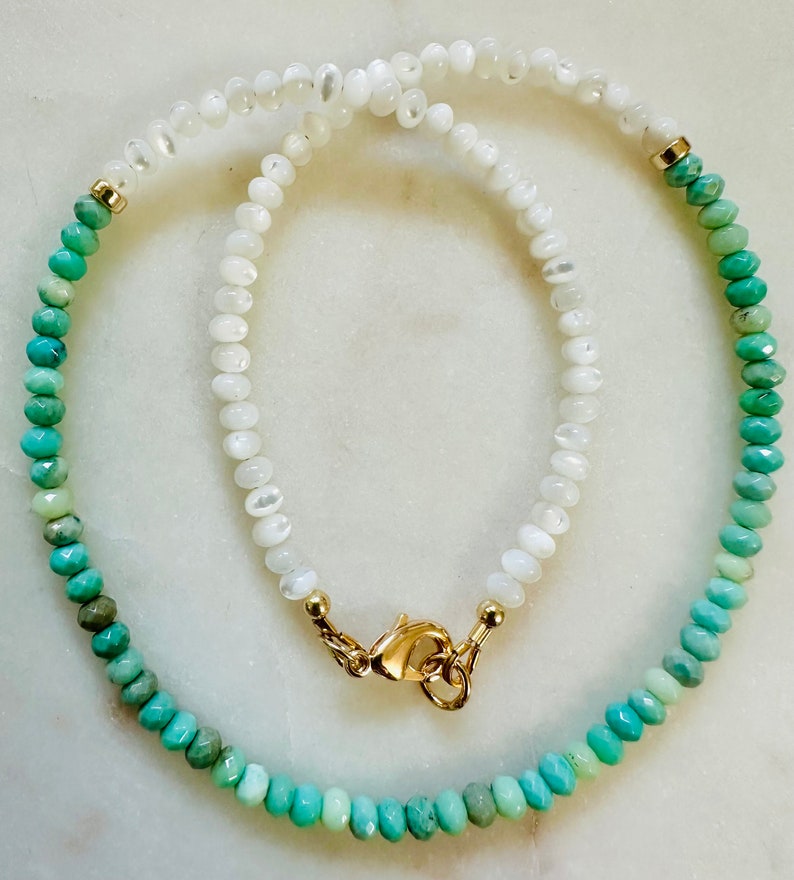 May include: A delicate necklace with a gradient of turquoise and white beads. The necklace is made with small, faceted beads and has a gold clasp.