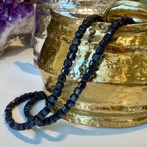 May include: A dark blue beaded necklace with square-shaped beads. The necklace is draped over a gold-colored decorative bowl. The background includes a purple crystal formation.