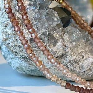 May include: Close-up of a delicate necklace featuring small, faceted beads in shades of peach and brown. The necklace is draped over a rough, gray stone, creating a contrast in textures. The beads appear to be strung together, forming a necklace.