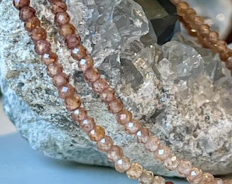 May include: Close-up of a delicate necklace featuring small, faceted beads in shades of peach and brown. The necklace is draped over a rough, gray stone, creating a contrast in textures. The beads appear to be strung together, forming a necklace.