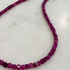 Puede incluir: Primer plano de un collar hecho de peque&ntilde;as piedras preciosas magenta oscuro, de corte cuadrado. Las piedras est&aacute;n ensartadas, creando un hilo flexible. El collar se muestra sobre una superficie blanca, similar al m&aacute;rmol.