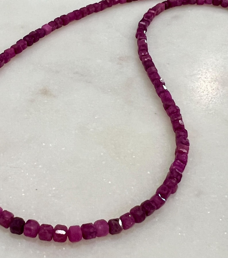 Puede incluir: Primer plano de un collar hecho de peque&ntilde;as piedras preciosas magenta oscuro, de corte cuadrado. Las piedras est&aacute;n ensartadas, creando un hilo flexible. El collar se muestra sobre una superficie blanca, similar al m&aacute;rmol.
