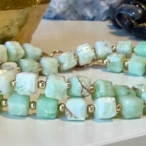 May include: A double-strand bracelet featuring light green, square-cut gemstones with gold-colored spacer beads. The gemstones have a natural, textured appearance. The bracelet is displayed on a white surface.