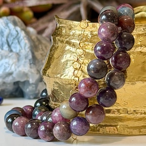 May include: A beaded bracelet featuring large, round beads in shades of purple, burgundy, and gray. The beads are strung on a gold-colored chain. The bracelet is displayed in front of a textured gold-colored vase.