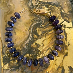May include: A beaded bracelet featuring oval-shaped, dark blue and purple gemstones. The bracelet is strung on a gold-colored chain with a matching clasp. The gemstones are evenly spaced around the wrist.