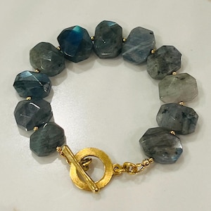May include: A gold tone bracelet with a toggle clasp features faceted labradorite beads in shades of blue, gray, and black.