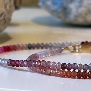 May include: A delicate bracelet composed of small, faceted beads in shades of pink, purple, and orange. The bracelet features a gold-colored clasp. The beads transition from light to dark hues, creating a gradient effect.