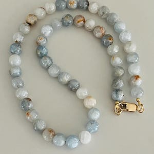 May include: A beaded necklace featuring round, light blue, white, and tan beads. The necklace has a gold-colored clasp. The beads are strung together to form a necklace.