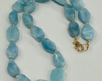 May include: A necklace featuring large, irregular-shaped aquamarine gemstones in varying shades of light blue. The stones are strung together with small gold-colored beads and a gold-colored clasp.