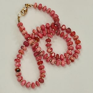 May include: A pink gemstone necklace with gold-colored accents. The necklace features small, irregularly shaped pink stones strung together with gold-colored beads. The clasp is gold-colored. The necklace is displayed on a white surface.