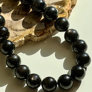 May include: A close-up of a black beaded necklace. The beads are round and shiny, reflecting light. The necklace is resting on a light-colored, textured surface. The beads appear to be approximately 1 inch in diameter.