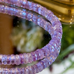 May include: Close-up of a delicate, faceted amethyst bead necklace. The light purple beads are strung together, reflecting light and creating a shimmering effect. The necklace is displayed on a reflective surface.