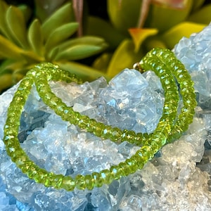 Peut inclure: Un collier délicat composé de petites perles de péridot vert clair, facettées. Le collier est posé sur un lit de cristaux bleu clair, avec un feuillage vert en arrière-plan. Les perles scintillent légèrement.