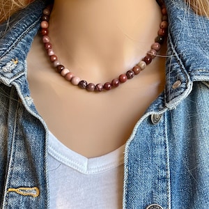 May include: A beaded necklace with a mix of red, pink, and brown tones. The necklace is composed of small, round beads and is worn against a white v-neck shirt and a denim jacket.