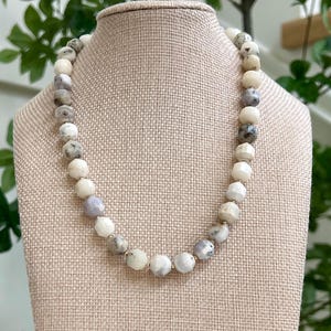 May include: A beaded necklace with a variety of round and faceted beads in shades of white, gray, and lavender. The necklace is displayed on a beige bust form, showcasing the jewelry's design and color variations.