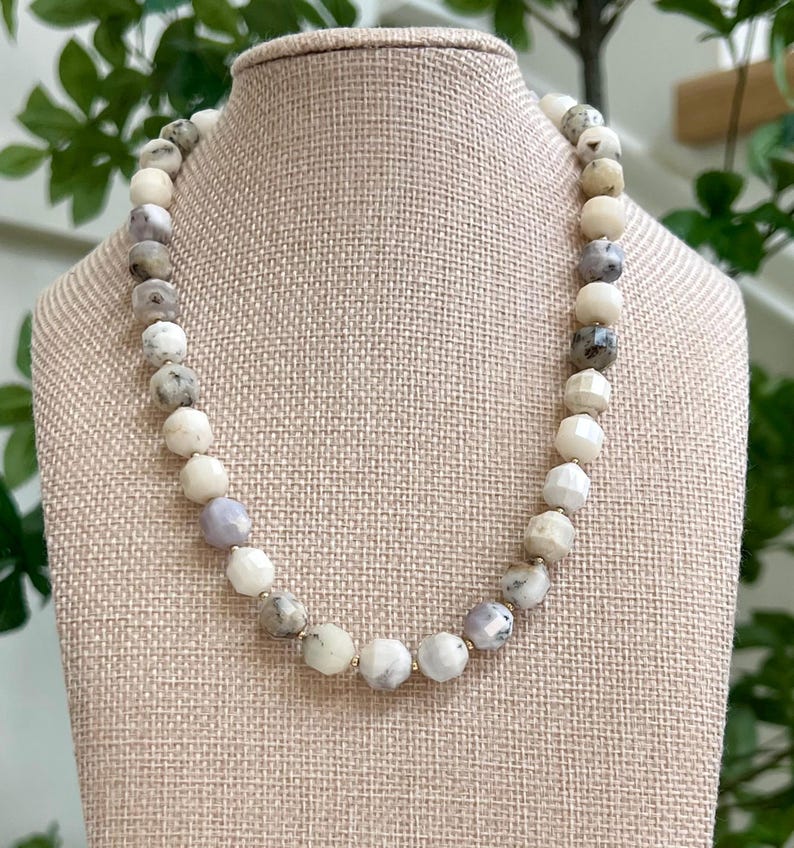 May include: A beaded necklace with a variety of round and faceted beads in shades of white, gray, and lavender. The necklace is displayed on a beige bust form, showcasing the jewelry's design and color variations.