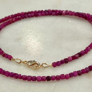 May include: A delicate necklace featuring small, faceted, deep pink ruby beads. The necklace is strung together with a gold-colored clasp. The beads are evenly sized and create a continuous loop, perfect for layering or wearing alone.