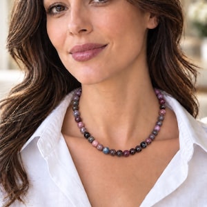 May include: A beaded necklace featuring round, polished stones in varying shades of deep red, purple, dark grey, and hints of blue. The beads are uniform in size and strung together to form a circular collar-style necklace worn around the neck.