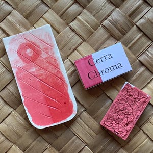 May include: Three pink watercolor paint pans, two are rectangular and one is a larger, oval-shaped pan. The rectangular pans have the text "Cerra Chroma" printed on them. The pans are on a woven, light brown surface.