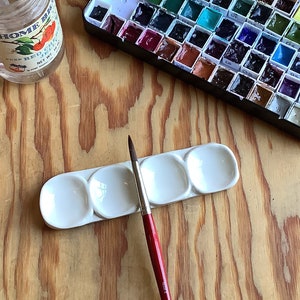 May include: A set of watercolor paints in a metal case, a white ceramic paint palette with four wells, and a red handled paintbrush on a wooden surface.