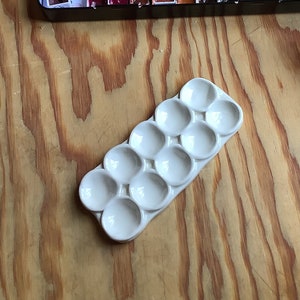 May include: A watercolor artist's setup on a wooden surface. A metal tin holds a variety of watercolor paints. A white ceramic palette, a jar of jelly, and a paintbrush are also present. The paints are in a range of colors.