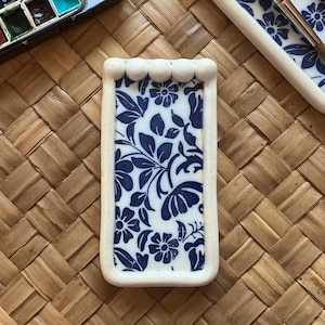 Puede incluir: Un juego de pintura de acuarela y dos soportes para pinceles con diseños florales azules. El juego de pintura está abierto, revelando una variedad de pinturas coloridas. Los soportes para pinceles son rectangulares con borde blanco y patrón floral azul.