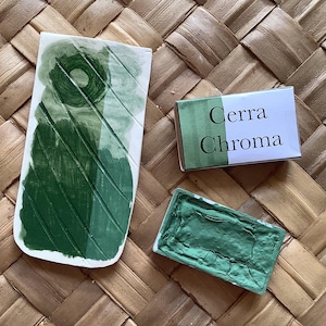 May include: Overhead view of art supplies: a rectangular watercolour paint palette, a small box with "Cerra Chroma" text, and a painted leaf shape, all in green hues. They are arranged on a woven, textured surface.