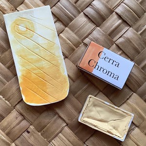 May include: A still life image of art supplies on a woven surface. A rectangular ceramic palette with yellow paint and diagonal lines, a small box labelled "Cerra Chroma", and a small paint pan filled with yellow paint.