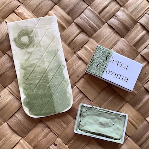 May include: A green watercolor paint pan with the brand name "Cerra Chroma" on the packaging. The paint pan is sitting on a woven surface.
