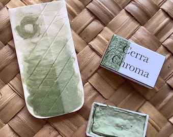 Può includere: Una pastiglia di acquerello verde con il nome del marchio "Cerra Chroma" sulla confezione. La pastiglia di acquerello è su una superficie intrecciata.