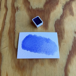 May include: A white plastic watercolor palette with a variety of colors, a small blue watercolor pan, a white paintbrush holder, a red paintbrush, and a blue watercolor wash on a white paper.