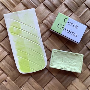 May include: A green watercolor paint pan with the brand name "Cerra Chroma" on the packaging. The paint pan is sitting on a woven mat.