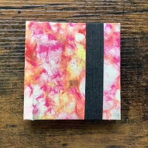 Puede incluir: Un pequeño cuaderno cuadrado con una cubierta de mármol rosa, naranja y amarillo. El cuaderno tiene un cierre de banda elástica negra.