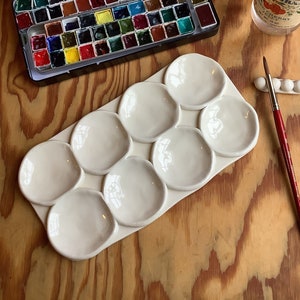 Peut inclure: Une palette en céramique blanche avec neuf puits ronds pour mélanger les aquarelles. La palette est à côté d'un jeu d'aquarelles dans une boîte en métal, un pot d'eau et un pinceau.