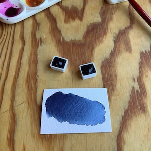 Puede incluir: Una configuración de artista de acuarela con una paleta blanca llena de varios colores, dos pastillas de acuarela cuadradas, un pincel y una muestra de pintura azul oscuro sobre papel blanco. Los elementos están dispuestos sobre una superficie de madera.